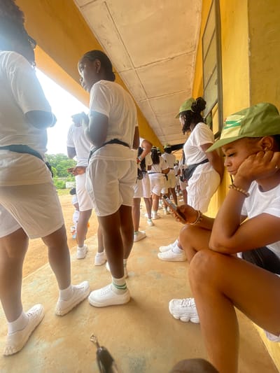 In Ise-Ekiti, Nigeria, a group of young people, likely participants in a youth corps program, are gathered outdoors under a yellow-painted building with a concrete overhang. The ground is a sandy, unpaved surface. Most individuals are wearing white t-shirts and white shorts, with some donning green caps.
In the foreground, a young woman is seated and looking at her mobile phone. She has dark skin, braided hair, and is wearing a white t-shirt, black shorts, white socks, and white sneakers. She has a beaded bracelet on her wrist. To her left, other individuals are standing, also in white attire. Some are looking forward, others are engaged in conversation. The scene appears to be during daylight hours, with natural light illuminating the area. The building's architecture suggests a functional, possibly institutional setting. The overall atmosphere is one of casual gathering and waiting.