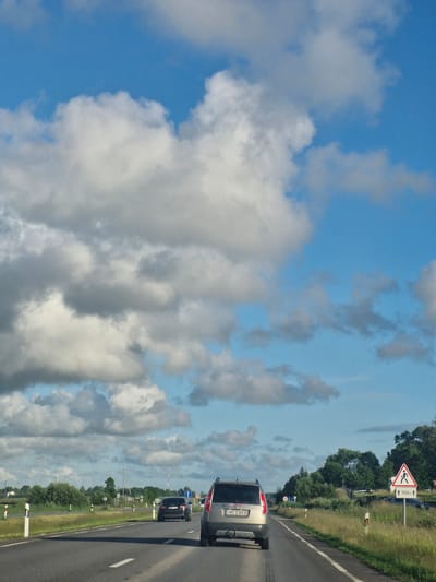 An over-the-shoulder shot captures a view from a moving vehicle traveling along a two-lane asphalt road. The road is flanked by a grassy verge and a line of trees. The sky above is a bright blue, dotted with fluffy white and gray clouds.

In the foreground, the rear of a silver SUV with the license plate "MCD 959" is visible. The vehicle has a tow hitch. Further ahead on the road, a black Audi sedan is in the left lane, its taillights illuminated. Beyond the Audi, traffic signs are visible, including a speed limit sign indicating "70" and a pedestrian crossing sign with an arrow indicating "700m."

The environment appears rural, with some houses and trees visible in the distance on the left side of the road. A cell tower can be seen in the distance on the right. The overall impression is of a clear, sunny day suitable for travel. The location is identified as Rimkai, Lithuania.