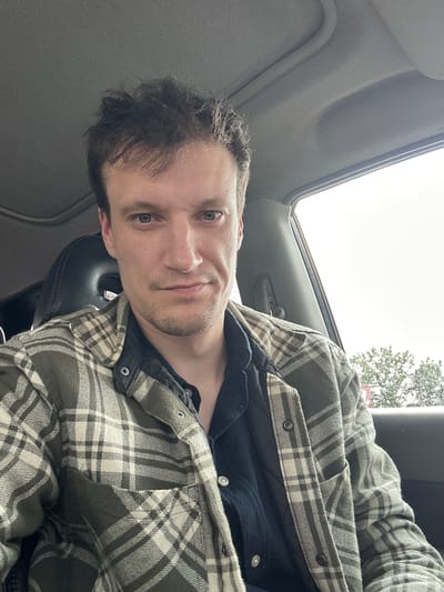 A man is sitting in the driver's seat of a car, taking a selfie. He has short, dark, messy hair and fair skin. He is wearing a dark collared shirt underneath a green and white plaid shirt jacket. The car interior is visible, including the headrest of the passenger seat and the car's ceiling. Outside the car window, green trees and a bright sky are visible, suggesting it is daytime. The man is looking directly at the camera with a neutral expression.