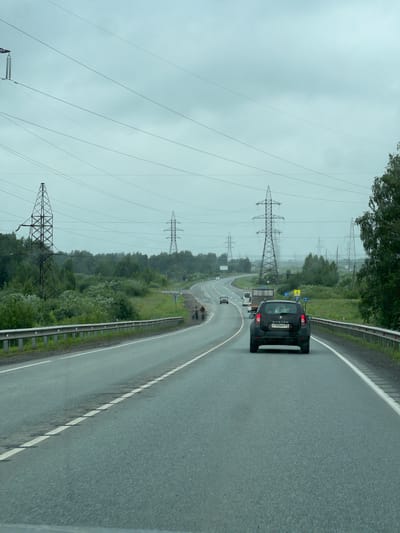 An overcast day frames a view from a vehicle traveling on a two-lane asphalt highway that curves to the right. The highway has white lane markings, with a dashed line dividing the lanes and a solid white line marking the shoulder. A silver metal guardrail runs along the right side of the road, with green vegetation beyond it. On the left side of the road, a continuous line of trees and bushes forms a dense barrier. In the distance, several large lattice-style power line towers are visible, interspersed with smaller towers, and a network of overhead power lines crisscrosses the sky.
In the foreground, a black SUV with license plate Y700AH is in the right lane, facing away from the viewer. To its left, a white truck is also traveling in the same direction, followed by a smaller white vehicle. Further ahead, a white delivery van is in the left lane. On the shoulder of the road, near a series of signs, two individuals are seen with bicycles, appearing to be on the side of the road. A flock of birds is in flight in the sky. The overall scene depicts a typical highway landscape in a rural or semi-rural area, characterized by infrastructure for power transmission and vehicular traffic under a cloudy sky. It's possible this scene is located near Krugloye, Russia, given the presence of power infrastructure and a highway.