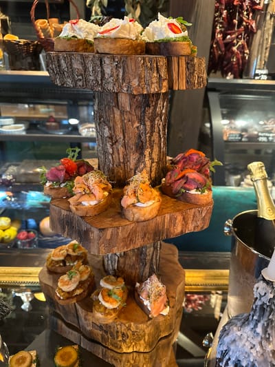 The image shows a food display, likely in a restaurant setting. A multi-tiered wooden structure made from rough-hewn logs holds several varieties of open-faced sandwiches or bruschetta. The sandwiches are topped with ingredients like whipped cream and strawberries, thinly sliced beef, salmon, and shrimp. A partially visible bottle of champagne sits above the display. The background contains reflections in glass and hints of the interior of the restaurant, including other food items and decorative elements. Given the city Новоглаголево, Russia, it is plausible that this is a local restaurant or food establishment.