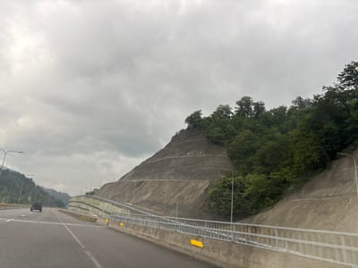 This is a wide-angle, eye-level, medium-distance shot of a highway in Tsakva, Georgia, on a cloudy day. The highway curves to the right, with multiple lanes visible. On the left side of the highway, there are forested hills and streetlights. On the right side, a steep, terraced, rocky hillside is reinforced with concrete structures and topped with dense green trees. A silver SUV is driving in the left lane of the highway, heading away from the viewer. A concrete barrier with a metal railing runs along the right side of the highway, separating it from the hillside. Yellow reflectors are attached to the railing. The sky is overcast and gray.