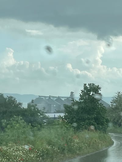 The image depicts a cloudy sky with a visible industrial structure in the background, likely a storage facility, possibly a grain elevator. There are trees and greenery along what appears to be a road or path on the right side of the image, with some red flowers interspersed. The presence of water droplets suggests rain or a wet surface. The photo was taken in Makariopolsko, Bulgaria.