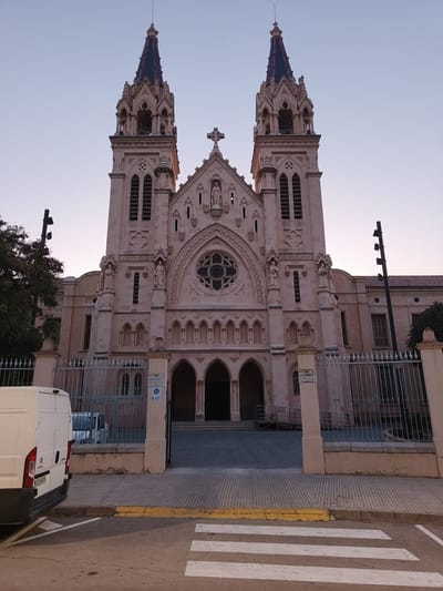 The image shows the exterior of a large, ornate church in Benirredrà, Spain, likely at dusk or dawn due to the soft lighting and clear sky. The church has a prominent facade with two tall bell towers topped with pointed, dark blue spires. The building is constructed of light-colored stone, possibly sandstone, and features Gothic architectural elements like pointed arches, intricate carvings, and decorative statues. A large rose window is visible above the main entrance.

In the foreground, a white delivery van is parked on the left side of the street, with its rear end facing the viewer. The van has a visible license plate. A pedestrian crossing with white stripes is painted on the asphalt in front of the van. The street is paved with grey cobblestones and bordered by a yellow curb.

A metal fence with decorative elements runs along the front of the church property, separating it from the sidewalk and street. Behind the fence, a paved area leads to the church entrance, which features a set of steps and a ramp. A few trees are visible on the left side of the image, behind the van and fence. The overall scene suggests a quiet, urban environment.