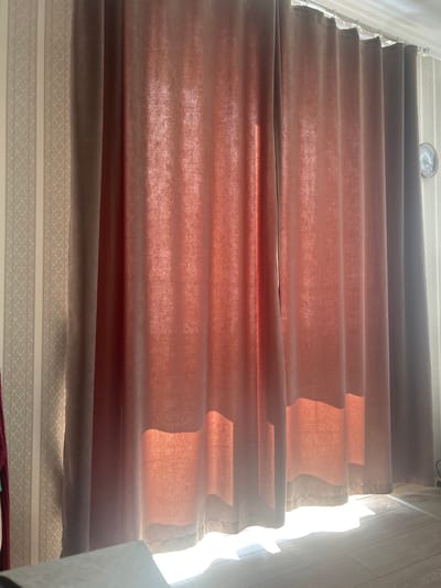 The image shows a close-up of a window with curtains in what appears to be a residential setting in Novayatura, Russia. The curtains are a dusky rose color and are partially drawn, allowing sunlight to filter through, creating a warm glow and casting shadows on the wall and floor. The wallpaper behind the curtains is a light neutral color with a subtle vertical striped pattern. The bottom of the curtains reaches the floor, which is visible as light-colored wood flooring. A small analog clock with a visible face and hands is mounted on the wall to the right of the curtains. No people or animals are visible in the image.