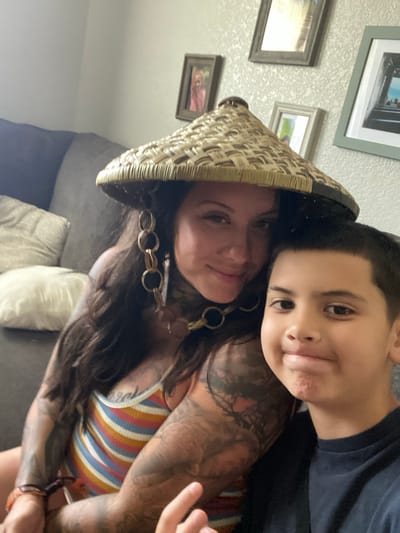 A woman with numerous tattoos and a conical hat adorned with large, golden hoop earrings is pictured with a young boy. They are seated on a couch, which is visible in the background. The woman is wearing a striped tank top, while the boy is in a dark t-shirt. The scene appears to be an indoor setting, possibly a living room, with framed pictures visible on the wall. The image was captured in Elizabeth City, United States.