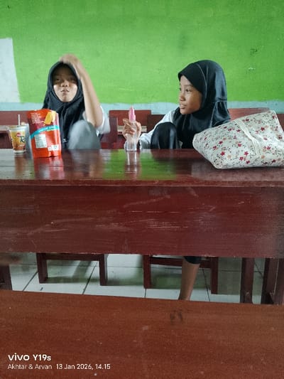 Two young girls wearing black hijabs sit at a wooden school desk in front of a green wall. The girl on the left has a bag of chips and a drink, while the girl on the right holds a small spray bottle and has a floral patterned bag on the desk. The scene is captured at a low angle, with another desk visible in the foreground. The picture includes a timestamp at the bottom.