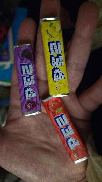 A hand holds three wrapped PEZ candies. The wrappers are colorful and printed with "PEZ" in bold, stylized letters. One is red and labeled "Strawberry" with a strawberry graphic. Another is yellow and labeled "Lemon" with a lemon graphic. The third is purple and features a character image along with a grape. The candies are positioned on the fingers and palm of the hand.