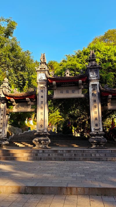 The image shows a stone gate structure with traditional Vietnamese architectural elements. The gate features pillars inscribed with Chinese characters, a tiled roof with ornate decorations, and an overhead plaque with more Chinese characters. The structure is situated at the base of a series of stone steps leading upwards, suggesting an entrance to a temple or sacred site. Lush green trees surround the gate, and a clear blue sky is visible above. The setting is likely in Lao Cai Ward, Vietnam, as indicated in the prompt.