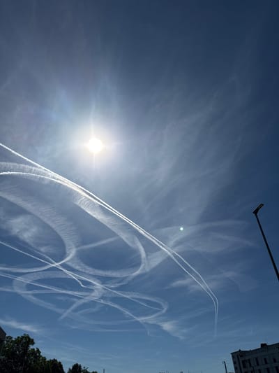 The image shows a bright, sunny sky with numerous contrails from airplanes forming an intricate pattern of white lines across the blue expanse. The sun is visible in the upper left portion of the frame, casting a bright glare. In the lower right corner, a modern street lamp is partially visible. At the very bottom of the image, the tops of green trees and parts of buildings are visible, suggesting an urban or suburban setting. The presence of contrails and buildings indicates a location likely in or near a city, such as Salach, Germany, which is known for its airspace being used by air traffic. There are no people or animals visible in the image. The scene is a snapshot of a clear day with active air traffic.