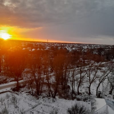 An aerial photograph depicts a snowy landscape at sunrise or sunset in Borodianka, Ukraine. The horizon on the left exhibits a bright orange and yellow sun, casting an intense glow across the lower left portion of the frame and illuminating the sky, which transitions to dark gray, heavy clouds towards the upper right. The middle ground features an extensive cityscape of residential buildings with snow-covered roofs. A prominent, slender industrial chimney or tower is visible on the distant horizon, slightly right of the center. The foreground and midground are covered in snow, featuring numerous bare deciduous trees with visible branches. A snow-dusted road or path curves into the scene on the far left. In the lower left, a fenced plot of land contains smaller, bare trees or shrubs. A portion of a snow-covered building roof, possibly corrugated metal, is visible in the lower right corner. No people or active subjects are visible in the scene.