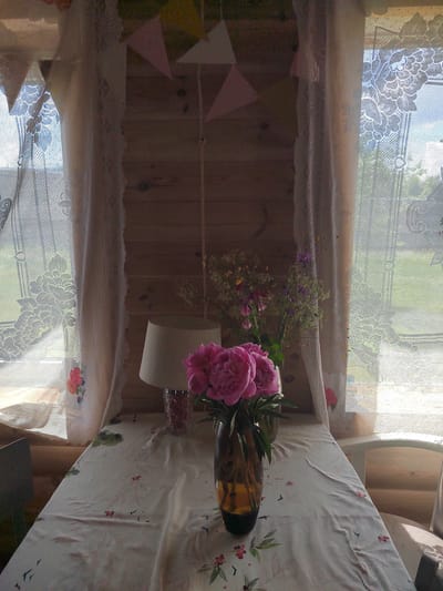 A dimly lit, rustic interior scene, possibly a sunroom or veranda, is captured. Sunlight streams through lacy white curtains on either side of a wooden-paneled wall. A light-colored tablecloth with a delicate floral and bird pattern covers a table in the foreground. On the table sits a dark amber vase filled with vibrant pink peonies, and to its right, a cluster of smaller wildflowers in a vase. A cream-colored lampshade is visible behind the peonies, placed atop a patterned glass jar. Bunting with pink and yellow triangular flags is strung across the top of the scene. The overall atmosphere suggests a tranquil, natural setting, perhaps in or near the Бярэзінскі сельскі Савет region of Belarus.