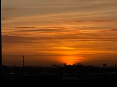 The image captures a vibrant sunrise or sunset over a Dutch urban or suburban landscape, specifically Maasdijk, Netherlands. The sky is dominated by striking warm hues of orange, golden yellow, and red, with horizontal, streaky clouds illuminated by the low-lying sun.
The sun itself is a brilliant orange-red semicircle, partially obscured by the distant skyline, glowing intensely on the horizon. Below the radiant sky, the landscape is rendered in dark silhouette. A prominent, tall communication tower stands on the left side of the mid-ground. The horizon line is composed of the dark silhouettes of various buildings and structures, including what appears to be a distinct multi-story building near the sun and a faint steeple or spire on the far right. A plume of what looks like smoke or steam rises from a point within the silhouetted buildings, just to the left of the sun.
The foreground is very dark, almost entirely black, suggesting rooftops or the top of a building from the vantage point of the photograph. No specific human activity or people are visible. The image conveys a calm, atmospheric scene at either dawn or dusk, characterized by the strong contrast between the luminous sky and the dark, silhouetted cityscape. There is no visible text in the image.