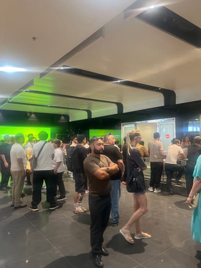 The image shows a group of people standing in a line, possibly waiting for something. They are inside a modern building with sleek architecture, featuring white walls and dark flooring. The lighting suggests it's daytime, but the interior setting makes it hard to determine the exact time or weather conditions.
The subjects are diverse in terms of age and gender, wearing casual clothing. Some individuals are looking forward, while others are engaged in conversation or looking down. The atmosphere appears relaxed, with no obvious signs of distress or urgency.
Notable details include a green illuminated wall or display in the background, which has some text that is difficult to read due to the image orientation and resolution. The overall scene conveys a sense of everyday life in a public space, likely a commercial or entertainment venue given the clean and contemporary design. The fact that it's Parakar, Armenia, provides a specific geographic context.