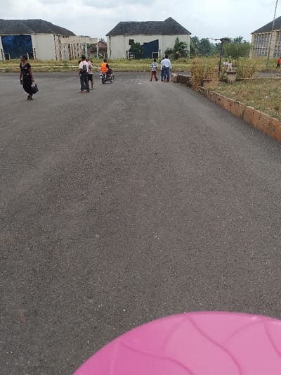 The image shows a paved area in Ogboko, Nigeria, with several buildings in the background. A woman in a dark outfit and carrying a bag is walking to the left. A group of people, including children, stand near the center. A motorcyclist is present. Two men stand further back. Some people stand in the distance. The buildings appear to be white with dark roofs. The foreground includes a blurred pink object. Overgrown vegetation is visible along the edges of the paved area. The sky is overcast.