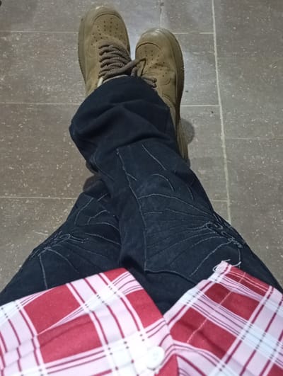 The image shows a close-up, high-angle view of a person's legs and feet, seated on a tiled floor. The person is wearing dark wash denim jeans with some embroidered detailing and a red and white checkered shirt. Their feet are clad in tan suede athletic shoes with dark laces. The floor appears to be made of large, rectangular gray tiles with white grout lines. The lighting suggests an indoor setting, likely during daylight hours given the natural light casting shadows. The scene does not provide specific clues to its exact location within Ukpor, Nigeria, beyond the general indication of a tiled floor and seated posture.