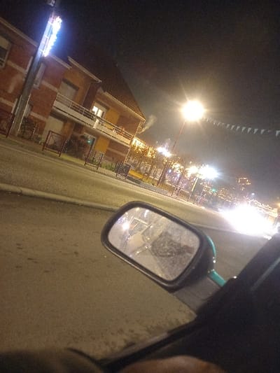 This nighttime image, taken in Sallaumines, France, captures a tilted street scene from the perspective of a vehicle. On the far right, part of a vehicle's dark body is visible, along with its side-view mirror, which reflects bright light sources and indistinct shapes from behind the camera.

The left side of the frame features a row of buildings. One prominent building has a reddish-brown facade with multiple windows and small balconies. Another building further along the street appears in shades of yellow and orange. Lights are visible from some of the building windows. A wisp of white smoke or vapor can be seen rising from behind one of these structures, possibly from a chimney.

The street is heavily illuminated by artificial lights. A very bright street lamp with a strong halo is prominent in the upper left section against the dark sky. Numerous strings of bright, festive lights, possibly holiday decorations, stretch across the scene above the buildings, contributing to a lively atmosphere. A string of small, triangular flags or bunting is also visible against the dark sky, suggesting a celebratory occasion. At the bottom left, an intensely bright, bluish light source, likely a commercial sign, adds further illumination.

No individuals are visible in the image. The time of day is clearly night, characterized by the dark sky and reliance on artificial light. No decipherable text is visible on any signs due to the brightness. The scene depicts an active, illuminated street within a town or city.