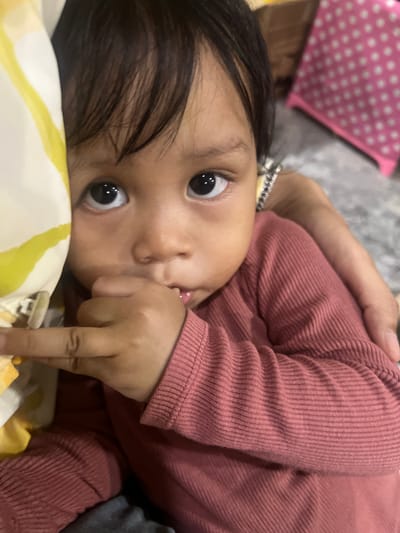 A close-up shot focuses on the face of a young child, who appears to be an infant or toddler. The child has dark hair, dark eyes, and is looking directly at the camera with a slightly curious expression. Their right hand is raised to their mouth, with fingers placed inside. The child is wearing a dark pink, ribbed long-sleeved shirt. To the left of the child's face, a portion of a yellow and white patterned fabric is visible, possibly a blanket or pillow. In the background, a blurred pink object with white polka dots is discernible, and to the right, an adult's arm is visible, lightly embracing the child. The overall setting appears to be indoors, with a neutral-colored, textured surface in the background, suggestive of a carpet or rug. The image was taken in Wedarijaksa, Indonesia.