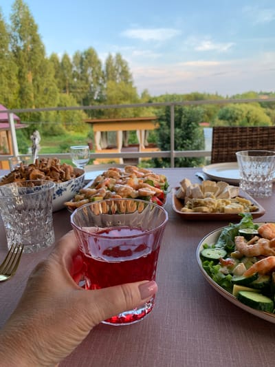 A close-up, first-person perspective shot shows a hand holding a glass of red liquid at a table set for a meal outdoors, overlooking a lush, green landscape with a body of water in the distance. The table is covered with a taupe tablecloth and features several dishes of food, including a large bowl of mushrooms, a platter of shrimp and salad, and a wooden plate with various cheeses. There are also two clear glasses, a gold fork, and a white plate on the table. The background shows a railing, trees, and a building structure by the water. The setting appears to be a tranquil, natural environment, potentially a dacha or countryside retreat, consistent with the rural location of Суроватиха, Russia. The lighting suggests it is either late afternoon or early evening.