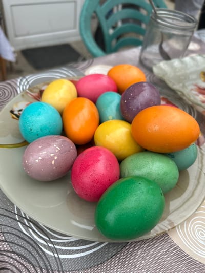 A top-down shot shows a plate piled high with brightly colored Easter eggs, mostly in shades of orange, purple, yellow, blue, red, and green. The plate sits on a tablecloth with a circular pattern in shades of gray, black, and white. A glass jar is slightly out of focus in the left of the image. The image does not provide enough information to ascertain if it was taken in Zlati Voyvoda, Bulgaria. There is no information provided in this image that can determine if people are in this scene or interacting.