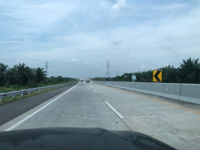 The image presents a front-facing view from inside a vehicle traveling on a multi-lane concrete highway under an overcast sky. The vehicle's dashboard occupies the lower portion of the frame.
The highway stretches into the distance, featuring clear white lane markings, including a solid white line on the left shoulder and dashed lines separating the lanes. On the left side of the road, there's a narrow asphalt shoulder alongside a silver metal guardrail equipped with red reflectors. Beyond the guardrail, lush green vegetation, dominated by numerous palm trees, lines the roadside.
On the right side of the highway, a tall, light grey concrete barrier runs parallel to the road. Attached to this barrier is a bright yellow chevron sign, pointing left, which typically indicates a sharp curve or bend in the road. Further along the right, behind the concrete barrier, a green highway sign with white text, including the number '27', is partially visible amidst dense palm trees and foliage.
Several electricity transmission towers are visible across the landscape on both sides of the highway and in the distant background, suggesting an area with developed infrastructure. A few other vehicles can be seen in the distance, also traveling along the highway. The sky is a uniform light blue-grey, covered with soft, scattered clouds, indicating a cloudy daytime setting. The environment suggests a tropical or subtropical region, consistent with the location context of Stabat, Indonesia. The scene depicts a routine journey along a well-maintained road.