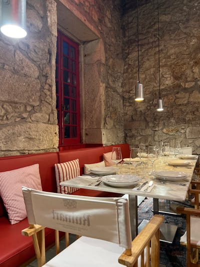 The image displays the interior of a dining establishment located in Caminha, Portugal. Rough-hewn stone walls define the space. On the left, a tall, narrow window with a dark red, multi-pane frame is visible. Below the window and extending across the wall is a continuous, upholstered bench seat in a solid red color. Several red and white striped cushions are placed on this bench. A long, rectangular dining table with a light-colored marble top occupies the foreground and mid-ground. This table is set for multiple diners, featuring white plates, silver cutlery, and various clear glass stemware, including wine and water glasses, arranged for at least eight settings. Three cylindrical, silver-toned pendant lights hang from the ceiling over the table, providing illumination. Director-style chairs with light wooden frames and white fabric backs are positioned around the table; the chair in the immediate foreground shows an inverted logo on its back, partially legible as "HUMM." A patterned rug is visible on the floor beneath the table. No individuals are present in the scene.