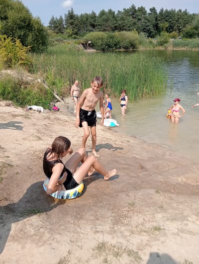 A group of people are at a lake on a sunny day. In the foreground, a young girl sits on the sandy shore in a black swimsuit, with an inflatable ring around her waist. A boy in black swim trunks stands near the water's edge, looking towards the girl. Several other people are in the shallow water: a man in black swim trunks, a young girl in a blue swimsuit, a woman in a dark bikini, and a woman in a pink bikini with a yellow inflatable. Another inflatable, shaped like a white and blue whale, is in the water. The background features tall reeds, trees, and a clear blue sky. The location appears to be a natural lake or reservoir. The weather is warm and sunny, typical for a summer day at a swimming spot. The mood seems relaxed and recreational, with people enjoying a day at the lake. There is no visible text in the image.