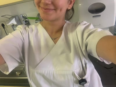 A female subject is depicted from the upper chest, smiling, with prominent dimples on her cheeks. She wears a light-colored, short-sleeved medical scrub top with a V-neck and a crossover front design. Small gold hoop earrings are visible in her earlobes, along with a thin silver-colored necklace featuring a small, reflective pendant. A black retractable ID badge reel is clipped to a pocket on her lower right side. The background environment suggests a clinical or institutional setting. On the left, a white wall-mounted dispenser, an electrical outlet with a black plug, and metallic fixtures are visible, alongside a green clip. On the right, a white wall-mounted Kimberly-Clark Professional paper towel dispenser with a circular indicator window is present, next to a partially visible blue object. The image provides no visual information to specify a precise location within the city of Nürtingen, Germany.