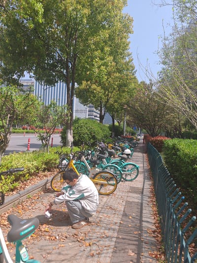 A man is crouching on a leaf-strewn sidewalk, adjacent to a row of parked bicycles, in what appears to be the Jianye District, China. The bicycles are predominantly light blue and yellow. The man is wearing a white jacket with green trim, blue jeans, and brown shoes. He is holding a small white object in his hand. Behind the bicycles is a green hedge and a dark green metal fence. In the background, there are trees, a multi-story building with vertical blue and white stripes, and a clear blue sky.