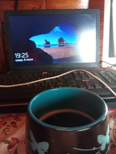 An overhead, medium shot shows a laptop with its screen displaying a desktop background of a beach scene at dusk. The time on the screen reads "19:25" and the date reads "среда, 8 июля". In the foreground, a dark brown mug with a teal rim is partially visible. The mug is decorated with light blue butterfly decals. The mug rests on a floral-patterned tablecloth. The laptop is on a wooden surface, and the background shows a wooden wall. The scene is indoors, and appears to be a personal workspace.