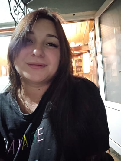 The image shows a young woman with long, dark hair, possibly in her late teens or early twenties. She is wearing a black sweatshirt with colorful embroidered lettering on the chest. The letters visible are "A", "L", "A", "K", and "E". The woman is looking at the camera with a slight smile and her eyes are dark. She has on light makeup, including blush and lipstick. A thin gold chain necklace is visible around her neck.

The photo appears to be a selfie taken indoors, possibly at night, as suggested by the artificial lighting in the background and the reflections on the window. The background shows a room with warm, orange-toned lighting and what looks like a window with a metal grate outside. The overall mood is casual and intimate, with the woman seeming relaxed and comfortable. The setting might be a home or a cafe. The image is slightly tilted, giving it a dynamic feel. The location context provided is Yevpatoriya, Ukraine.