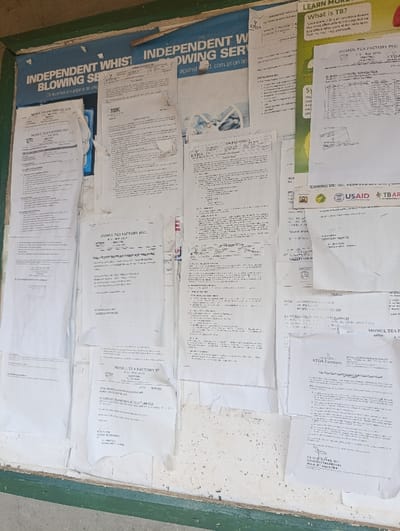 The image captures a weathered public information board in Kaplelwo, Kenya, covered with numerous white papers and posters. The board, possibly a wall, features a dark green frame at the bottom and a prominent blue banner across the top. The documents are tacked or pasted haphazardly, showing significant signs of wear, including crumpling, tearing, and curling edges, indicative of prolonged exposure.
The top blue banner displays "INDEPENDENT WHIST BLOWING SER" and "INDEPENDENT W BLOWING SER against corruption ar", suggesting an anti-corruption whistleblower service, with a partially visible phrase below, "Chagua hifadhi superna na uw...". To its right, a poster titled "LEARN MORE" educates on "What is TB?" (Tuberculosis).
A majority of the documents are official notices and announcements from "MOMUL TEA FACTORY PLC" and "MOMUL TEA FACTORY CO. LTD," frequently mentioning "KTDA" (Kenya Tea Development Agency). These papers include various lists, tables, and paragraphs of text, likely pertaining to factory operations, schedules, or regulations. Other visible entities include "USAID" and "TBAR" under a "CONTACT US" section. The board serves as a central point for disseminating important community information related to the local tea industry, public health, and governance. No people or specific activities are depicted; the scene is a static display of these public communications.