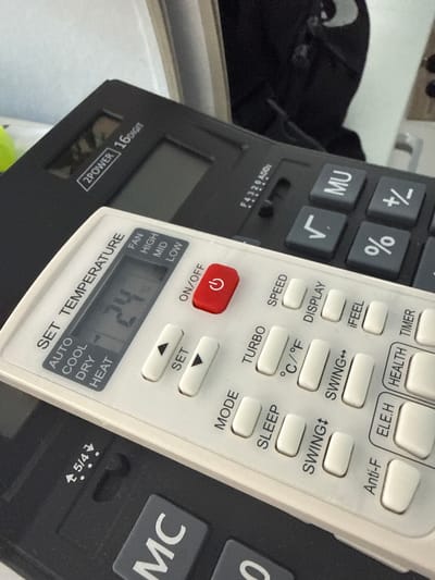 This media file shows a close-up view of two electronic devices, likely a calculator and a remote control, placed on a surface. The calculator is black with gray buttons and a digital display. Visible text on the calculator includes "MU", "%", "+/-", and "2POWER 16DIGIT". The remote control is white with numerous buttons and a small LCD screen. Text on the remote control includes "ON/OFF", "SET", "MODE", "TURBO", "SPEED", "DISPLAY", "İFEEL", "SWING", "°C/°F", "Anti-F", "ELE.H", "HEALTH TIMER", and "SET TEMPERATURE". The remote's screen displays "24" and indications for fan speed (FAN, HIGH, MID, LOW) and operating modes (AUTO, COOL, DRY, HEAT). The scene appears to be indoors, with diffused lighting. There are no people visible, and the setting lacks specific environmental cues that would indicate a particular time of day, weather, or precise location beyond the general context of electronic devices. The image's focus is on the details of the buttons and displays of the devices.