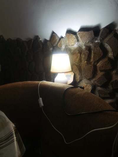 A bedside lamp illuminates a textured, stone-like wall behind a couch in a dimly lit room. The lamp has a white base and a cream-colored shade. A white cord with an inline switch trails from the lamp, lying across the couch. The couch is covered in a brown fabric with a subtle geometric pattern. Part of another object with a striped pattern is visible on the left side of the frame. The scene appears to be a residential interior in Lomets, Bulgaria.