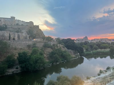 The image depicts a view of the city of Toledo, Spain, at dusk. In the foreground, a wide, calm river reflects the muted colors of the sky. The riverbanks are lined with lush green trees and vegetation. On the left side of the image, a steep hillside is covered with ancient stone fortifications and buildings, appearing to be part of the historic city walls and structures. Scattered streetlights are visible along pathways on the hillside. In the background, the Toledo skyline is visible, dominated by a large, domed cathedral, characteristic of Spanish cities. The sky displays a gradient of blues, purples, and pinks, indicating the setting sun, with some scattered clouds. The overall impression is one of historical grandeur and natural beauty during the twilight hours.