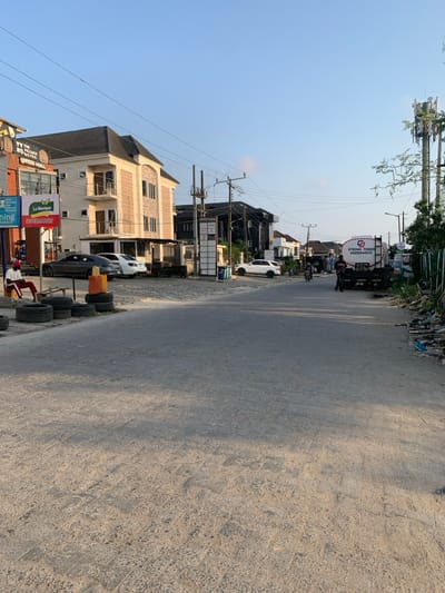 The image depicts a street scene in Ikota, Nigeria, under a clear, light blue sky with bright, even illumination suggesting daylight. The paved road, exhibiting some wear and tire marks, extends into the distance, narrowing slightly.
On the left side of the foreground, a stack of approximately ten black tires is present on a rough, unpaved shoulder. Adjacent to the tires, an orange barrier or drum is visible. A male individual, wearing a red long-sleeved shirt, dark trousers, and a white cap, is seated on a wooden bench or structure behind the tires, facing towards the road.
Further down the left side, a multi-story building with a dark roof and light-colored walls houses businesses, identifiable by signage including "THE VILLAGE BISTRO," "EXPRESS MALL," "La Monique," and "BIZEMARKET." Several vehicles are parked on the paved area in front of this building: a dark grey sedan, a white sedan, and a white SUV. A multi-panel signpost stands nearby.
Further down the street on the right side, a dark-colored multi-story building is visible, with a white SUV parked in front of it. On the right side of the road, a black flatbed truck with a large white tank-like container is parked. The tank displays a "G STRONG TOWER" logo and the phone number "070556644510." A male individual, dressed in a dark shirt and dark trousers, stands near the rear of this truck.
In the center of the road, a motorcycle with a single rider is in motion, heading away from the viewer. Utility poles with numerous overhead wires line both sides of the street. On the far right, a tall cellular communication tower is prominent. The right shoulder of the road appears unpaved and contains scattered debris and refuse. Sparse trees and vegetation are visible along the right side and in the background.