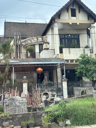 The image depicts a multi-story house in Quảng Trị Ward, Vietnam. The house has a tiled roof, white and beige exterior walls, and dark-colored windows. There is a covered porch area at the front of the house with a red and yellow lantern hanging from it. The yard in front of the house is overgrown with grass and features a black metal fence. Several stone sculptures and potted plants are visible in the foreground, along with stacks of wood and other building materials. The overall scene suggests a residential property with some decorative elements and a somewhat unkempt yard.
