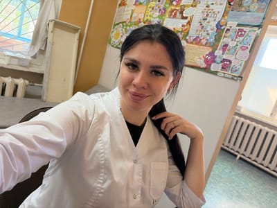 A young woman with dark hair and heavy makeup, wearing a white lab coat, smiles at the camera. She is seated in what appears to be a classroom or office, indicated by the colorful educational posters on the wall behind her and a radiator visible in the background. A window with a grate is also present. The overall impression is of a candid selfie taken indoors. The location is not specified in the image, and therefore cannot be determined.
