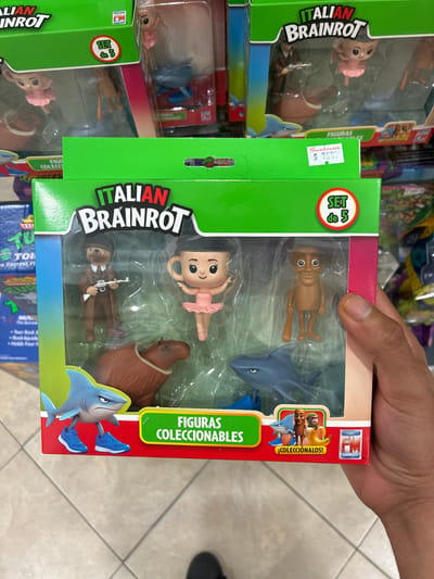A person holds a toy set in what appears to be a store. The toy set is labeled "Italian Brainrot," and contains five figures: a wooden-looking person, a ballerina doll, a figure of a bear with a gun, a shark, and a seal. The packaging also displays the text "Figuras Coleccionables" and "¡Colecciónalos!" Additionally, a "Fotoroma" logo can be seen on the packaging. Another toy set is visible in the background, suggesting the location is a store. A price tag of "Sanborns $7682" is visible, suggesting the location is a Sanborns store in Coatzacoalcos, Mexico. The background shows more toys on shelves and a tiled floor.