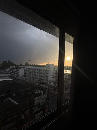 This image captures a view through a dirty window of a residential area in Surat Thani City Municipality, Thailand, at sunset. The sky is a dramatic mix of dark, stormy clouds on the left and a bright, sunlit area on the right, with the sun appearing as a hazy orb near the horizon. The cityscape below includes several multi-story apartment buildings with white facades and dark window frames, as well as lower-rise buildings with dark roofs. Electrical wires and poles are visible throughout the scene. The foreground is dominated by the dark interior of the room from which the photo is taken, with the window frame creating a strong border. There are no people or discernible animals visible in the image.