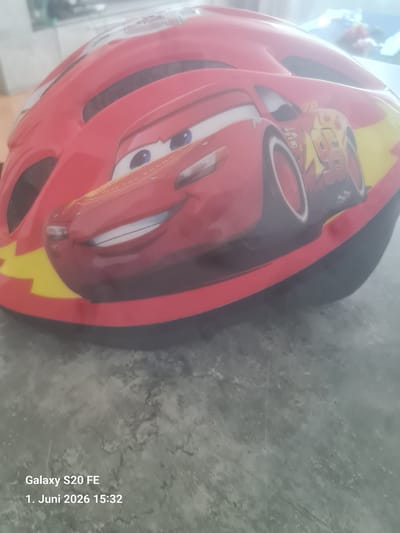 A close-up, slightly blurred photograph taken indoors on a surface, shows a red bicycle helmet with a cartoon image of Lightning McQueen from the movie "Cars". The helmet is also decorated with yellow lightning bolts. The background is an out-of-focus, light-colored surface. Text on the bottom right corner indicates "Galaxy S20 FE, 1. Juni 2026 15:32". There is no evidence to suggest the location is Bingen am Rhein, Germany.