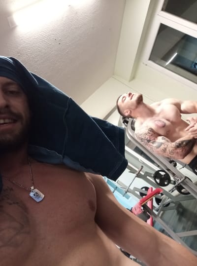 Two shirtless men are pictured in a gym. One man is close to the camera, wearing a dark blue fabric as a head covering and a dog tag necklace. His chest is visible, revealing a tattoo. The other man is further away, lying on a weight bench with his face turned upward and tattoos on his arm and chest. Weights and gym equipment are visible in the background. There is a window in the upper right corner.