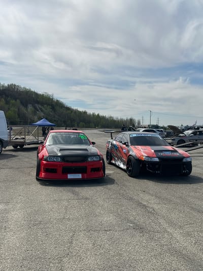The image shows two modified race cars parked on a grey asphalt surface, likely a staging area or paddock at a racetrack. The sky is overcast with predominantly white clouds. In the background, there is a line of trees on a slight hill, and some utility poles are visible. The red car in the foreground features a black hood and the number "16" on the side window. It also displays a "Systeme electric" logo on the front bumper. The second car, positioned slightly behind the red one, has a black and red livery with Cyrillic script. A few other vehicles are visible in the distance. Based on information on this image, the scene is likely at the Sadovoe racetrack in Russia.