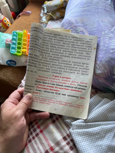 The image displays a close-up, overhead view of an open book with Cyrillic text, resting on a patterned fabric. The book's pages are slightly yellowed with age. To the left of the book, a colorful pop-it toy is visible, as well as part of a package labeled "AURA". A hand, with visible fingernails, is holding the book open. The background consists of a light-colored, patterned fabric with a floral or abstract design in shades of purple and white. The scene appears to be indoors, possibly in a domestic setting. There are no specific landmarks or details that would indicate the location of Кемуль, Russia.