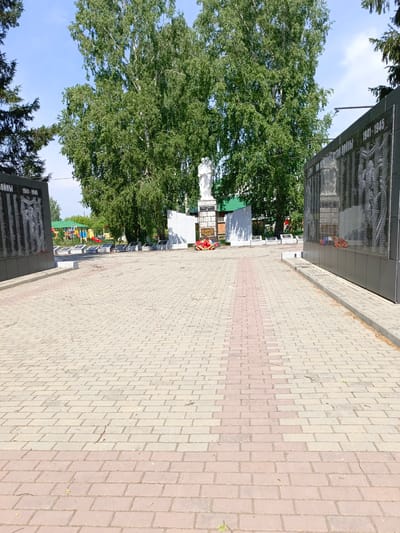 This is a ground-level, wide-angle photograph taken outdoors in Zalesovo, Russia, on a sunny day. The foreground is dominated by a paved walkway made of light tan and pink rectangular bricks laid in a repeating pattern. A path of pink bricks runs down the center of the walkway, extending into the middle ground.
In the middle ground, on the left side of the image, is a dark, multi-panel monument. The panels appear to be made of polished black stone, inscribed with rows of names and dates, with one panel showing a bas-relief image of figures. The inscription "1941-1945" is visible on one of the panels. To the left of this monument, a dark green coniferous tree is partially visible. In front of the monument, a white statue of two figures stands on a pedestal. A wreath of red flowers is placed at the base of the statue. Further back, a white, rectangular structure with a green roof is visible. Beyond that, a colorful playground with slides and swings can be seen.
On the right side of the image, a large, dark, multi-panel monument similar to the one on the left is present. It is also made of polished black stone with inscribed names and dates, and bas-relief images. The inscription "1941-1945" is clearly visible. Red floral wreaths are placed at the base of this monument as well.
The background features a large number of mature deciduous trees with dense green foliage, suggesting it is summertime. The sky is blue with some wispy white clouds. The overall scene depicts a war memorial with a surrounding park-like environment.