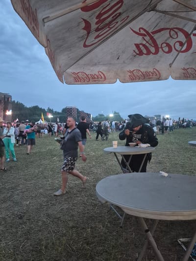 The image depicts an outdoor event, possibly a music festival, taking place in a grassy field under an overcast sky. A large umbrella with the Coca-Cola logo is prominently featured in the foreground, partially obscuring the sky. In the middle ground, a person wearing a black leather jacket and a wide-brimmed hat is seated at a round table, eating from a bowl. They are holding a spoon and appear to be focused on their meal. To the left of this individual, other people are visible, some walking and others standing. In the background, a crowd of people is gathered, with stage structures and trees suggesting a festival setting. The ground is covered in dry grass and some debris. The overall atmosphere appears casual and lively. The image is believed to have been taken in Lagunovo, Russia.