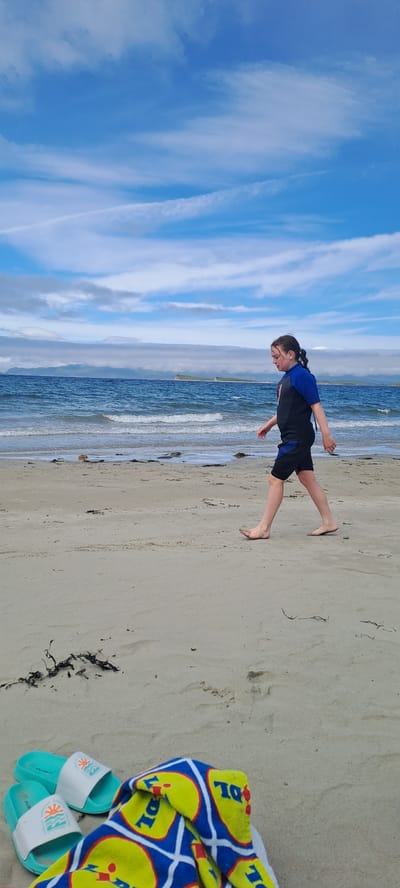 A young person wearing a blue and black wetsuit walks on a sandy beach. The person is walking from left to right across the frame. The ocean is to the left of the person, with waves breaking on the shore. In the background, there are islands and mountains. The sky is blue with white clouds. In the foreground, on the sand, there are two turquoise and white sandals and a folded towel with a yellow and blue pattern. The scene appears to be on Pier Road, Ireland.