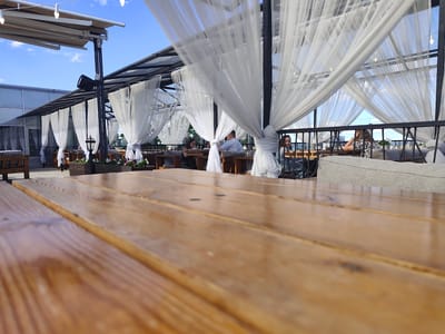 The image captures a scene at an outdoor rooftop restaurant or lounge in Petrozavodsk, Russia, during the daytime. The foreground is dominated by a close-up, low-angle view of a polished wooden table, its surface reflecting the light.

In the background, a covered seating area is visible, characterized by a metal frame structure draped with flowing white sheer curtains that create a sense of airy elegance. The curtains billow gently, suggesting a light breeze. Several people are seated at tables within this area, engaged in dining or conversation. One woman in the mid-ground on the left is looking down at her table, while a man in a light blue shirt sits opposite her, also looking down. Further to the right, another woman with dark hair looks towards the camera, and a woman with blonde hair in a patterned top is seated with her hand on her head, appearing relaxed.

The setting suggests a pleasant day, with a clear blue sky visible above the covered structure. The overall ambiance is relaxed and social, typical of a leisurely outing at a restaurant or cafe. The presence of tables, chairs, and people dining indicates that this is likely a functional establishment. The architecture of the roof and the lighting fixtures suggest a modern design. No visible text is present in the image.