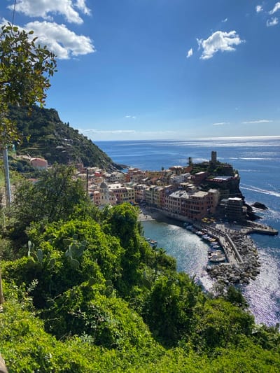 This high-angle image captures the coastal town of Vernazza, Italy, under a clear blue sky with scattered white clouds. The town is nestled into a steep, verdant hillside on the left, which is covered in dense green foliage, including discernible prickly pear cacti in the foreground. The Mediterranean Sea, a deep blue with sun-dappled reflections, extends to the horizon on the right.
The central feature is Vernazza's small, crescent-shaped harbor, protected by a stone breakwater and an extending concrete pier. Densely packed, multi-story buildings with colorful pastel facades (terracotta, pink, yellow, orange) line the harbor and climb the surrounding slopes. The Church of Santa Margherita d'Antiochia, identifiable by its octagonal bell tower and dome, is prominent near the waterfront. Perched on a rocky promontory overlooking the sea to the right is the Doria Castle, marked by its visible stone tower.
Numerous small boats are moored within the harbor and alongside the pier. A sandy/pebble beach is visible at the head of the harbor. A significant number of small figures representing people are dispersed across the beach, along the waterfront promenade, and densely populating the pier, indicating active pedestrian movement. Faint wakes from distant watercraft are visible in the open sea. The overall scene is brightly illuminated by direct sunlight, indicating daytime conditions.
