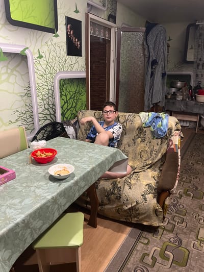 A boy with glasses is lying on a couch in a room. He is wearing a blue shirt and shorts. He is holding a gray object, possibly a book or tablet. A table covered with a green patterned tablecloth stands in front of the couch. On the table are two bowls, one red and one white, containing food. A black fan is near the table. The walls are decorated with green and white patterned wallpaper. Pictures hang on the wall. A striped garment hangs behind the couch. A patterned rug covers part of the floor. This image appears to be taken indoors. The location, Vesyoloye, Russia, can not be determined from the image.